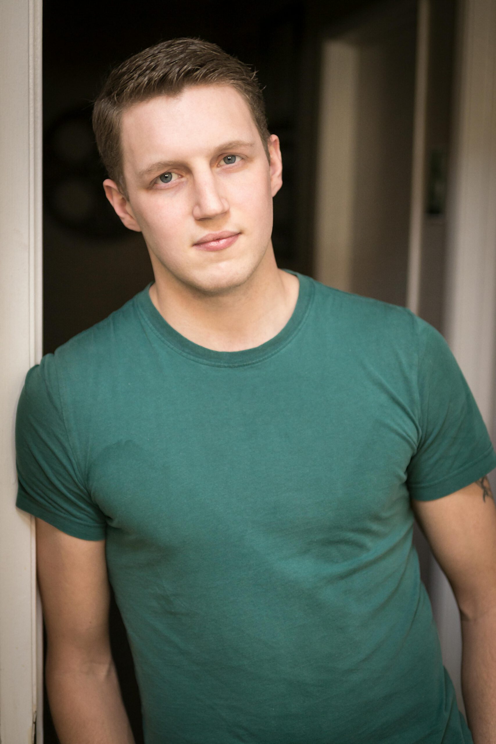 Relaxed portrait of a young man in a green shirt standing in a doorway.